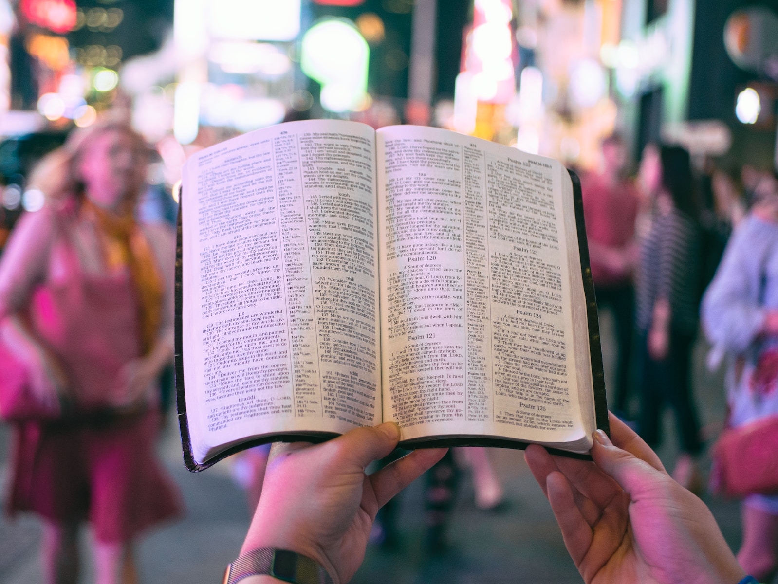 person holding bible on road with people walking on sidewalk beside buildings during nighttime